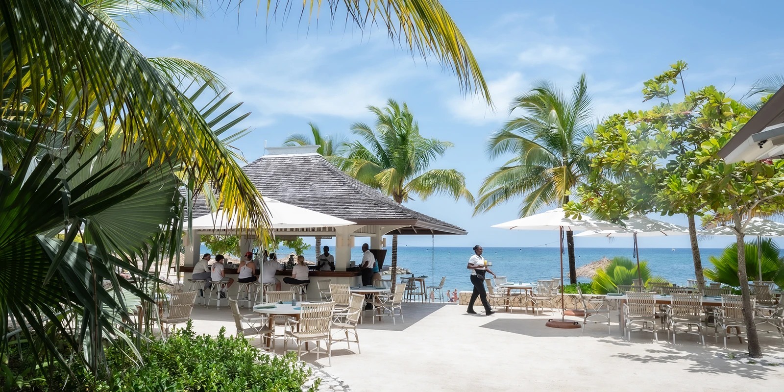 The beach bar terrace at midday — tables under palms, the sea just beyond