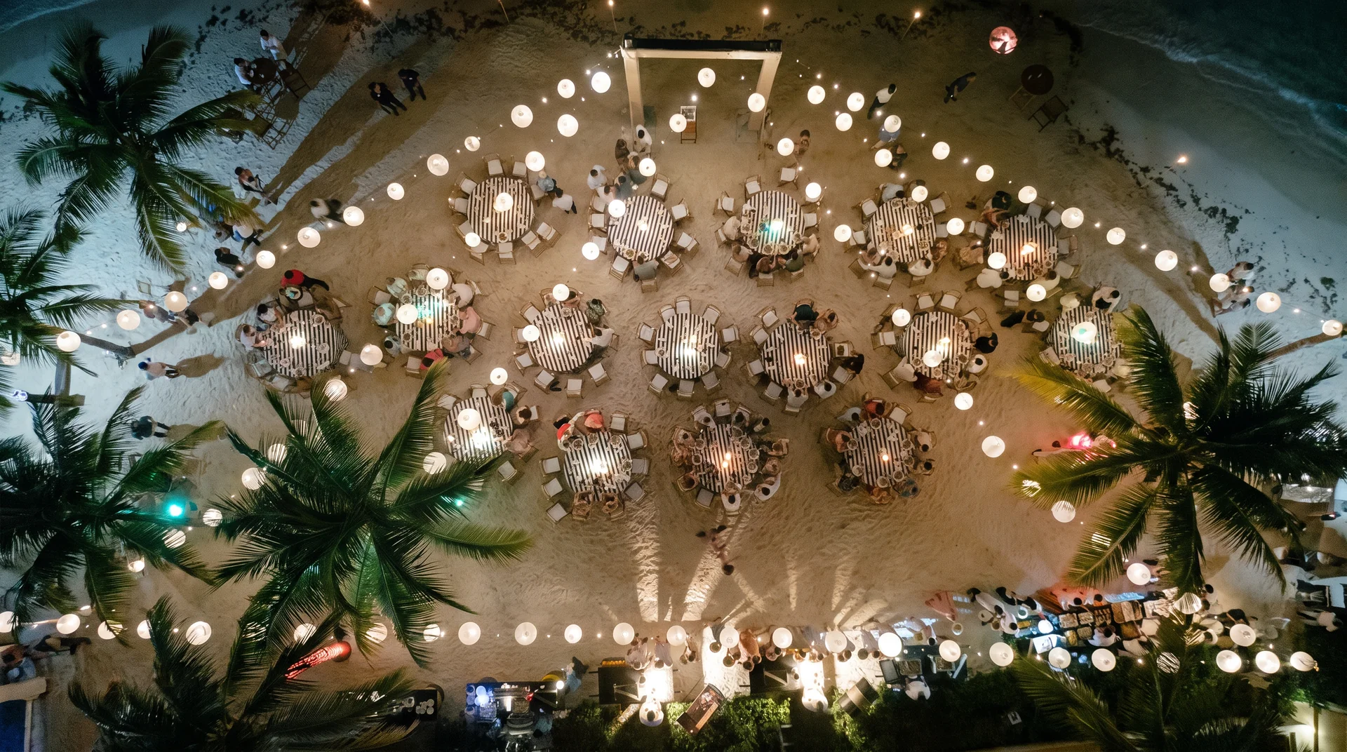 Lantern-lit dinner tables on the sand at night, seen from above and ringed by palms